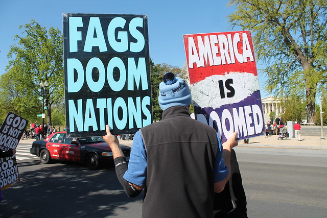 rear view of a protester holding signs which say "Fags Doom Nations" and "America Is Doomed"